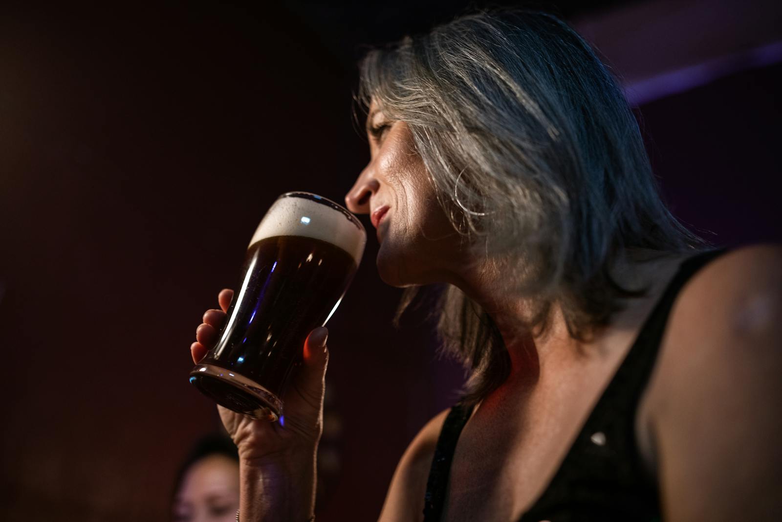 A mature woman sipping beer at a dimly lit bar, capturing a moment of relaxation and enjoyment.